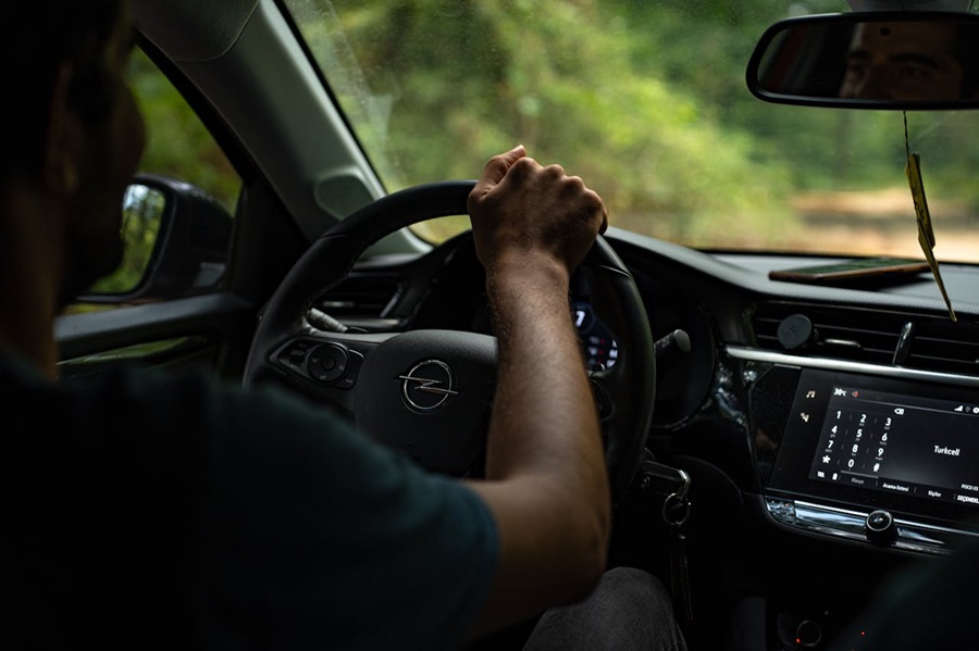 A man is driving without road tax through a forested area, gripping the steering wheel of a modern Opel car.