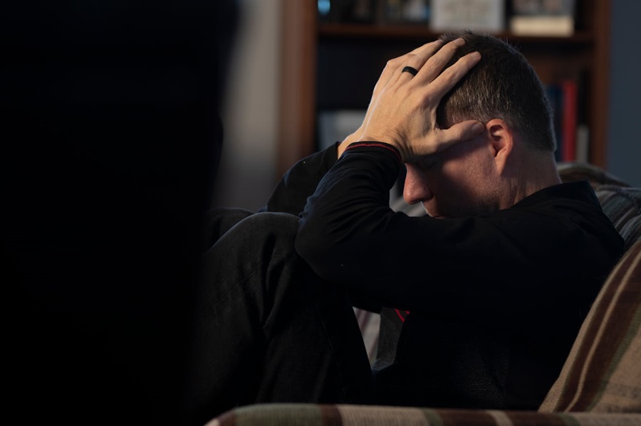 A man victim of domestic violence sits on a sofa with his head in his hands, looking distressed.