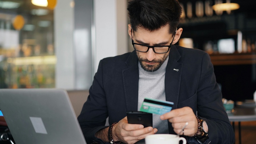 A man sitting at a table looking at his phone and holding a credit card, filing a complaint against a bank